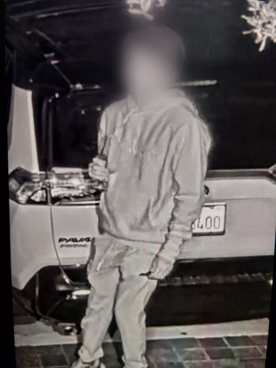 A person in a hoodie and pants stands in front of a parked SUV in a dimly lit area, holding an object in their right hand.